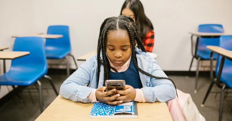 A young girl uses her smartphone while seated in a classroom environment, focused and engaged.