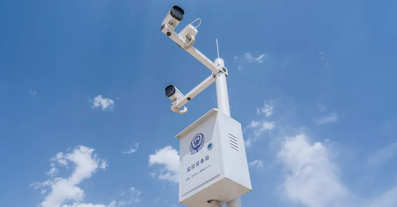 White security cameras mounted on a pole against a bright blue sky with fluffy clouds.