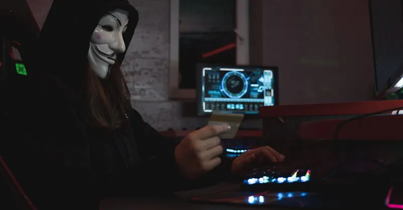 Masked hacker with credit card at computer, symbolizing cybercrime and anonymity.