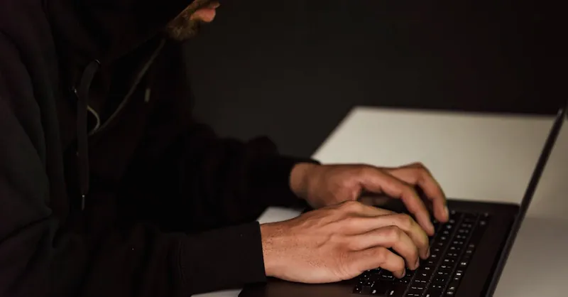 A person in a hoodie working on a laptop in a dimly lit room, representing cybersecurity themes.